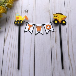 May include: A cake topper featuring two black sticks connected by a white banner with the orange letters T, W, and O. On the left is a yellow and orange excavator, and on the right is a yellow and brown dump truck, both attached to the sticks.