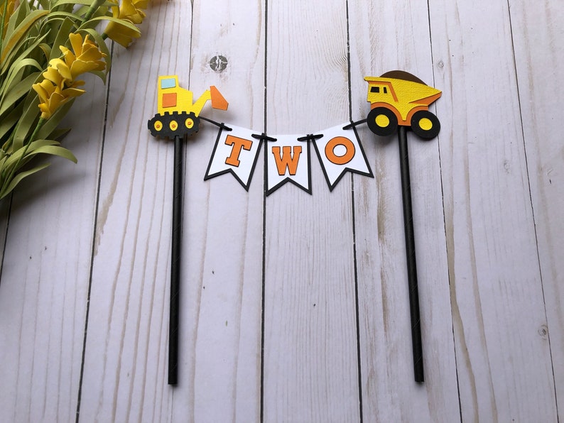 May include: A cake topper featuring two black sticks connected by a white banner with the orange letters T, W, and O. On the left is a yellow and orange excavator, and on the right is a yellow and brown dump truck, both attached to the sticks.