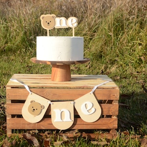 May include: A white cake with a brown bear and the word "one" on top. The cake is on a wooden cake stand and is sitting on a wooden crate with a brown bear and the word "one" banner.