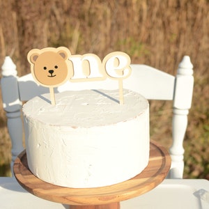 May include: A white cake with a brown bear and "one" cake topper on a wooden cake stand.