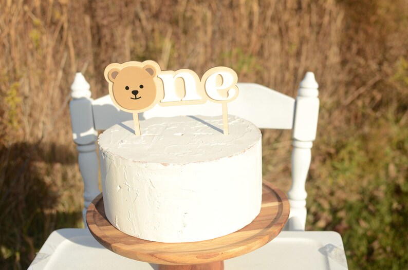 May include: A white cake with a brown bear and "one" cake topper on a wooden cake stand.