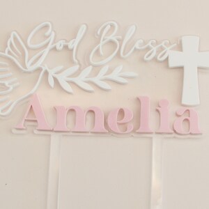 May include: A clear acrylic cake topper with the words "God Bless Amelia" in white and pink. The design includes a white dove, olive branch, and a white cross, suitable for a baptism or christening.
