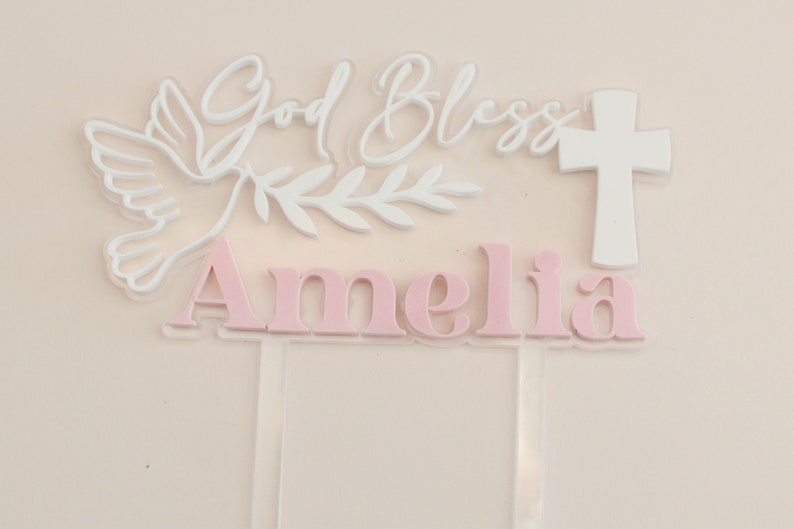 May include: A clear acrylic cake topper with the words "God Bless Amelia" in white and pink. The design includes a white dove, olive branch, and a white cross, suitable for a baptism or christening.