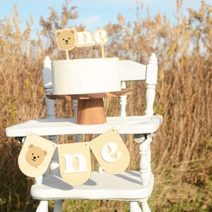 May include: A white cake with a brown bear topper and the word "one" on a white high chair with a brown banner that says "one" with a brown bear on the left side. The high chair is in front of a field of tall brown grass.