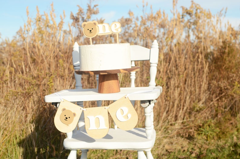 May include: A white cake with a brown bear topper and the word "one" on a white high chair with a brown banner that says "one" with a brown bear on the left side. The high chair is in front of a field of tall brown grass.