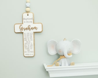 May include: A white and natural wood cross wall hanging featuring the text "Baptized 2.19.23", the name "Graham", and the scripture "Numbers 6:24-26", suspended by a string with light-colored beads, next to a small white elephant figurine.