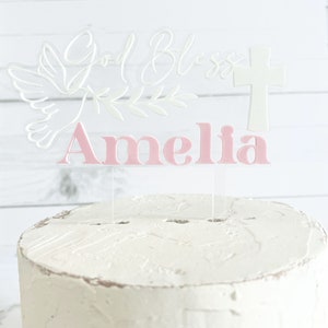 May include: A clear acrylic cake topper with the words "God Bless" in cursive, a dove, and a cross. The name "Amelia" is in pink block letters. The topper is inserted into a white frosted cake.