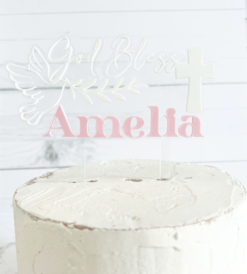 May include: A clear acrylic cake topper with the words "God Bless" in cursive, a dove, and a cross. The name "Amelia" is in pink block letters. The topper is inserted into a white frosted cake.