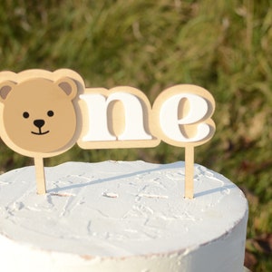 May include: A white cake with a brown bear face and the word "one" in white letters on wooden cake toppers.