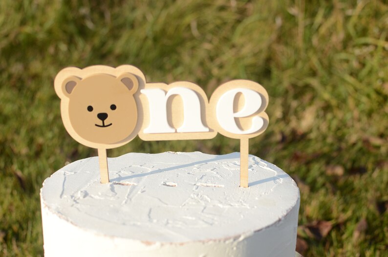 May include: A white cake with a brown bear face and the word "one" in white letters on wooden cake toppers.