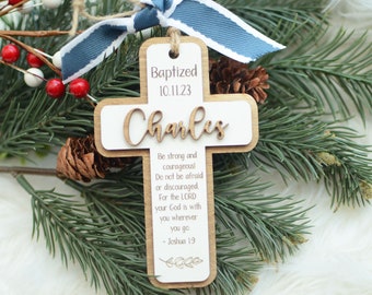 May include: A wooden cross ornament with a white background. The cross is decorated with the text "Baptized 10.11.23" and the name "Charles". The cross also features a Bible verse from Joshua 19: "Be strong and courageous! Do not be afraid or discouraged For the LORD your God is with you wherever you go - Joshua 19"