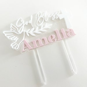 May include: A clear acrylic cake topper with the name "Amelia" in pink letters. The topper features a white dove, olive branch, cross, and the words "God Bless" in white script. The topper has two clear stakes.