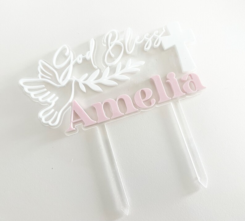 May include: A clear acrylic cake topper with the name "Amelia" in pink letters. The topper features a white dove, olive branch, cross, and the words "God Bless" in white script. The topper has two clear stakes.