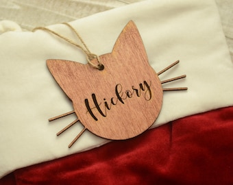 Puede incluir: Un adorno de Navidad de madera con forma de gato con el nombre "Hickory" grabado en él. El adorno está colgado de una media blanca y roja.