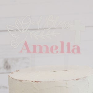 May include: A clear acrylic cake topper with the words "God Bless" and a cross. A dove and olive branch are etched into the acrylic. The name "Amelia" is in pink letters. The topper is placed on a white frosted cake.