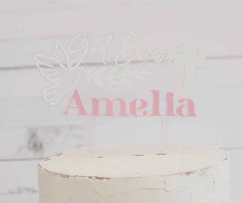 May include: A clear acrylic cake topper with the words "God Bless" and a cross. A dove and olive branch are etched into the acrylic. The name "Amelia" is in pink letters. The topper is placed on a white frosted cake.