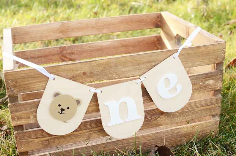 May include: A wooden crate with a white banner that says "one" in white letters. The banner has a brown bear face on one end.