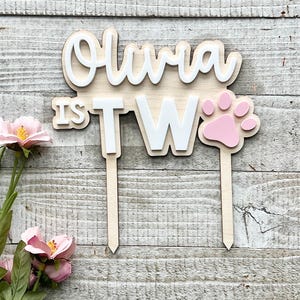 May include: A wooden cake topper with the name "Olivia" in white, layered over a light wood base. The text "IS TW" is also in white, with a pink paw print. Two wooden stakes extend from the bottom.