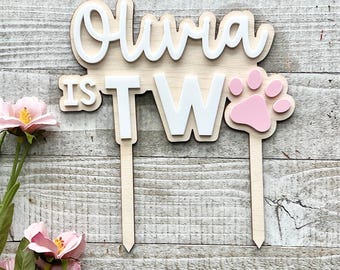 May include: A wooden cake topper with the name "Olivia" in white, layered over a light wood base. The text "IS TW" is also in white, with a pink paw print. Two wooden stakes extend from the bottom.