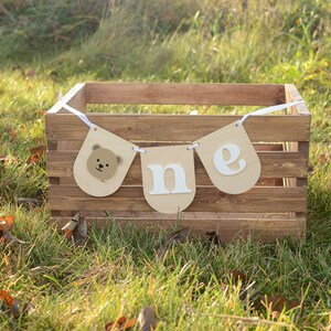 May include: A brown wooden crate with a white ribbon and a banner that says "one" with a bear face on the left side. The banner is hanging from the crate.