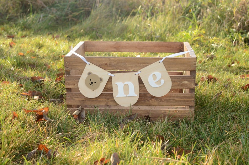 May include: A brown wooden crate with a white ribbon and a banner that says "one" with a bear face on the left side. The banner is hanging from the crate.