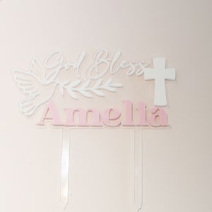 May include: A cake topper with the words "God Bless Amelia" in white and pink. The design includes a dove, olive branch, and a cross. The topper is made of clear acrylic with a pink background.