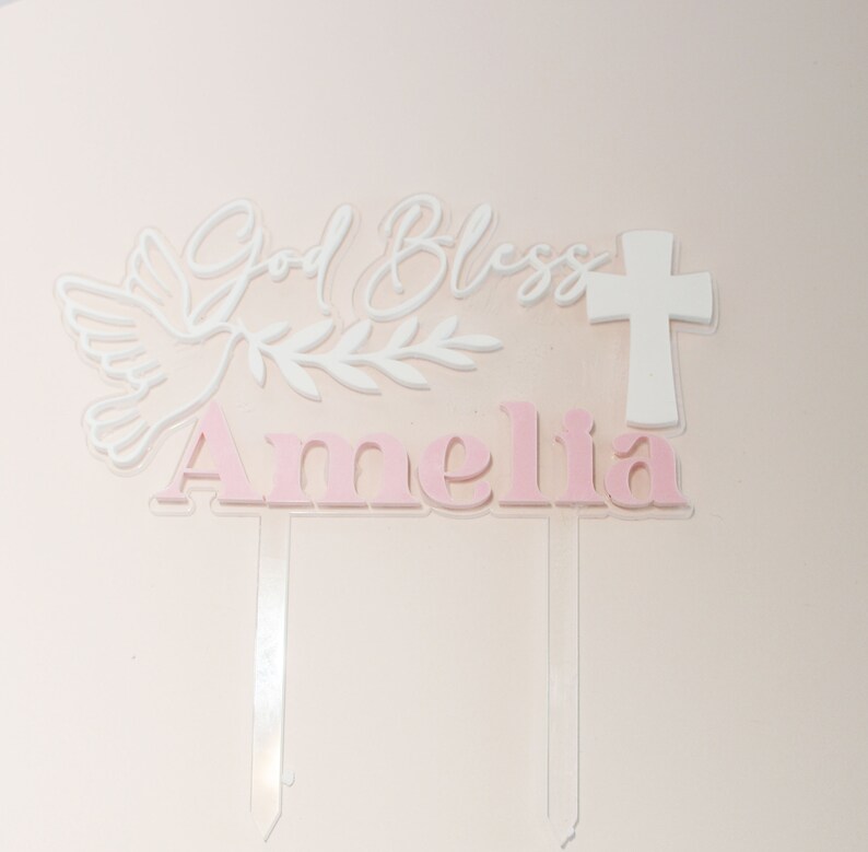 May include: A cake topper with the words "God Bless Amelia" in white and pink. The design includes a dove, olive branch, and a cross. The topper is made of clear acrylic with a pink background.