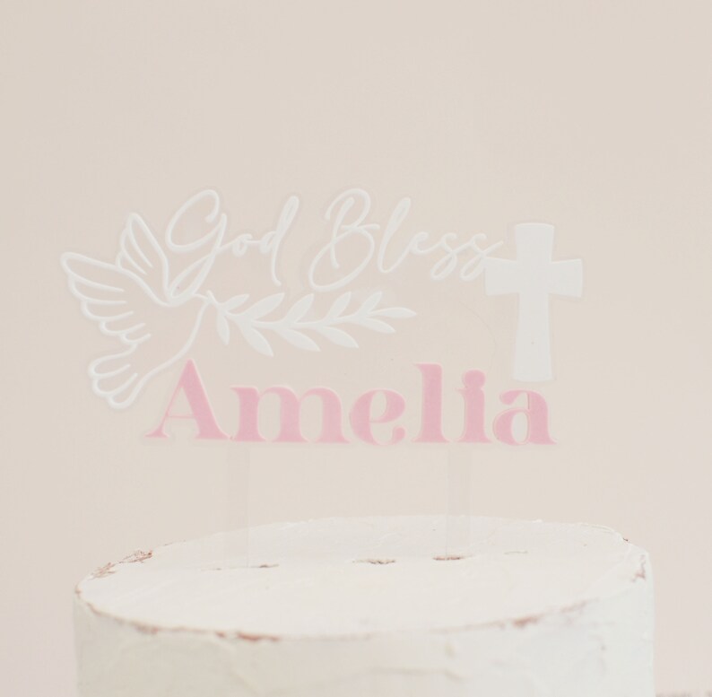 May include: A clear acrylic cake topper with the words "God Bless Amelia" in white and pink. The design includes a white dove, olive branch, and a cross, suitable for a baptism or christening celebration.