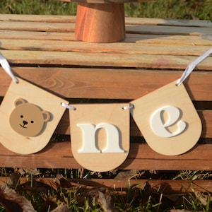 May include: A wooden banner with white letters spelling "one" and a brown bear face on the first flag. The banner is hanging on a wooden crate.