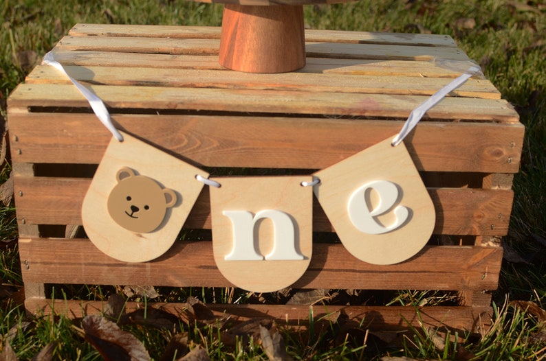 May include: A wooden banner with white letters spelling "one" and a brown bear face on the first flag. The banner is hanging on a wooden crate.