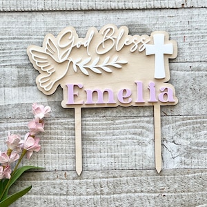 May include: A wooden cake topper with the words "God Bless" and the name "Emelia" in pink. The topper features a white dove, a cross, and a sprig of leaves. The background is a light wood grain.