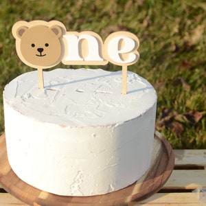May include: A white cake with a brown bear and "one" cake topper on a wooden cake stand.