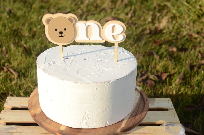 May include: A white cake with a brown bear and "one" cake topper on a wooden cake stand.