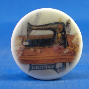 Peut inclure: Un bouton en céramique blanc et rond orné d'une illustration détaillée d'une machine à coudre Singer noire vintage sur une table en bois. La machine présente des détails dorés et le mot SINGER est clairement visible sur sa base.