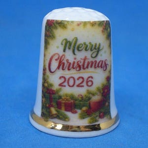 May include: A white ceramic thimble featuring a gold-trimmed base and a dimpled top. The front displays a festive Christmas scene with green pine branches, red gift boxes, and a mug, with the text Merry Christmas 2026 printed in red and green.