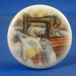 Può includere: Un pomello rotondo per mobili in ceramica bianca con un'illustrazione stampata di una macchina da cucire nera vintage su un tavolo di legno, con un colorato patchwork drappeggiato in primo piano, che mostra quadrati di tessuto rossi, gialli e blu.