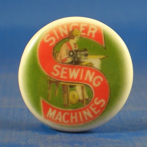 Może przedstawiać: Okrągła biała ceramiczna gałka z dużą czerwoną literą S i nadrukowanym białym napisem Singer Sewing Machines. Wzór zawiera ilustrację osoby obsługującej maszynę do szycia na zielonym tle z błyszczącym wykończeniem.