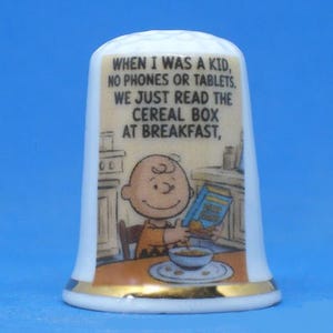 May include: A white ceramic thimble with a gold rim featuring a cartoon image of Charlie Brown reading a cereal box. The thimble has the text: "WHEN I WAS A KID, NO PHONES OR TABLETS. WE JUST READ THE CEREAL BOX AT BREAKFAST,"