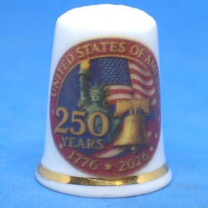 May include: White ceramic thimble with a gold band at the base. The thimble features a colorful design with the text "UNITED STATES OF AMERICA" and "250 YEARS 1776 2026" along with images of the Statue of Liberty, a waving American flag, and the Liberty Bell.