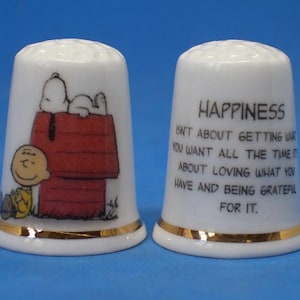 May include: Two white ceramic thimbles with gold trim. One thimble features Snoopy on his doghouse with Charlie Brown. The other thimble has the word "HAPPINESS" and a quote. Both thimbles are on a blue background.