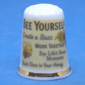 May include: A ceramic thimble in white and yellow, with gold edging. It features a bee and sunflower design, and the text "BEE YOURSELF, Create a Buzz, WORK TOGETHER, Sip Life's Sweet Moments, Stick Close to Your Honey."