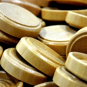 May include: Close-up of a pile of wooden checkers. The checkers are light brown with a circular shape and a slightly raised edge. The image is well-lit, highlighting the natural wood grain and texture of the checkers, suggesting a classic board game.