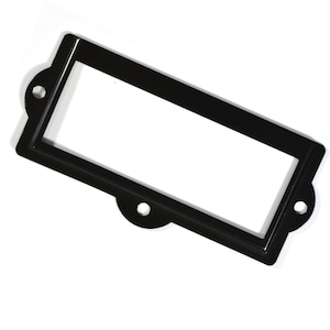 May include: Black metal label holder with rounded corners and two holes on each side. The holder has a rectangular opening for inserting a label.