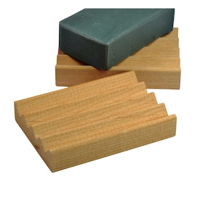 May include: A wooden soap dish with a stepped design, holding a rectangular bar of green soap. The soap dish is made of light-colored wood, and the soap has a smooth, solid appearance. The design allows for drainage.