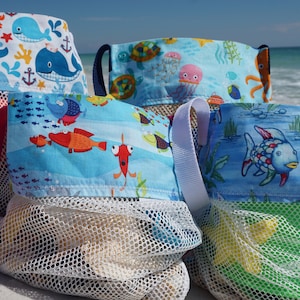 May include: Four colourful mesh beach bags with different sea creature designs. The bags are filled with toys and are sitting on a white sandy beach.