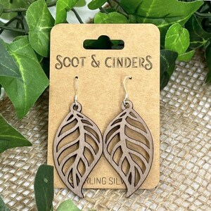 Könnte beinhalten: Holz-Blatt-Ohrringe mit Sterling-Silber-Haken, präsentiert auf einer braunen Karte mit dem Text "Scot & Cinders". Die Ohrringe haben ein detailliertes Blattdesign mit ausgeschnittenen Adern. Der Hintergrund zeigt grüne Blätter und Jute.