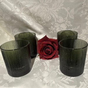 May include: Four olive green textured glass tumblers. The glasses have a vertical lined pattern and are arranged with a red rose on a white floral patterned tablecloth. The glasses are cylindrical and have a slightly angled rim.