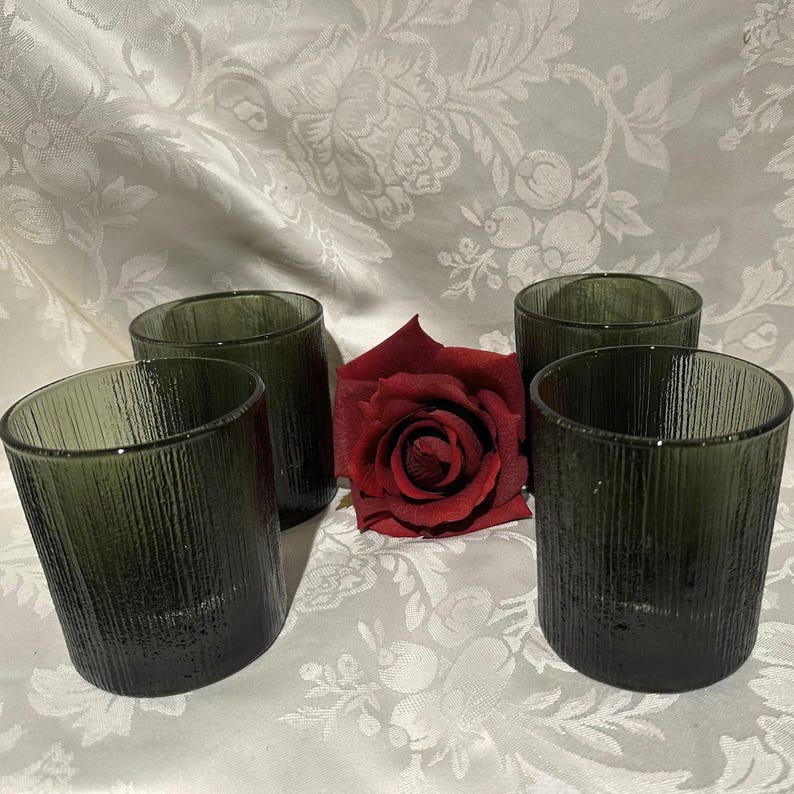 May include: Four olive green textured glass tumblers. The glasses have a vertical lined pattern and are arranged with a red rose on a white floral patterned tablecloth. The glasses are cylindrical and have a slightly angled rim.
