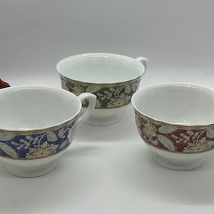 May include: Three white teacups with floral designs in blue, green, and burgundy. Each cup has a handle and sits on a small pedestal base. The floral patterns include leaves and flowers in shades of gold and cream.