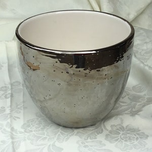 May include: A silver-colored ceramic bowl with a textured surface and a dark rim. The interior is white. The bowl has a wide opening and a rounded shape, suitable for various uses.